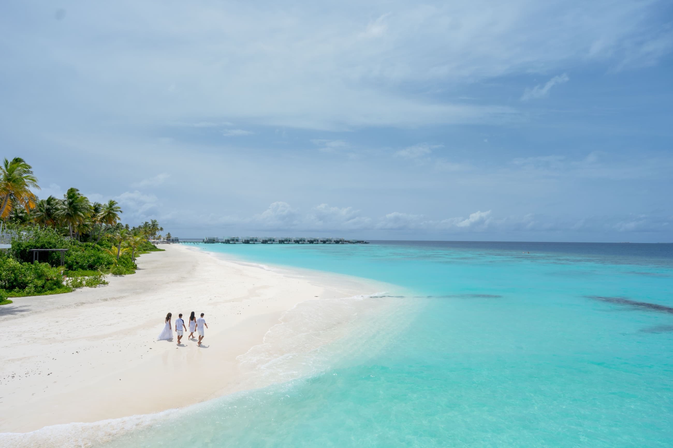 A group walking along a white sand beach past overwater villas