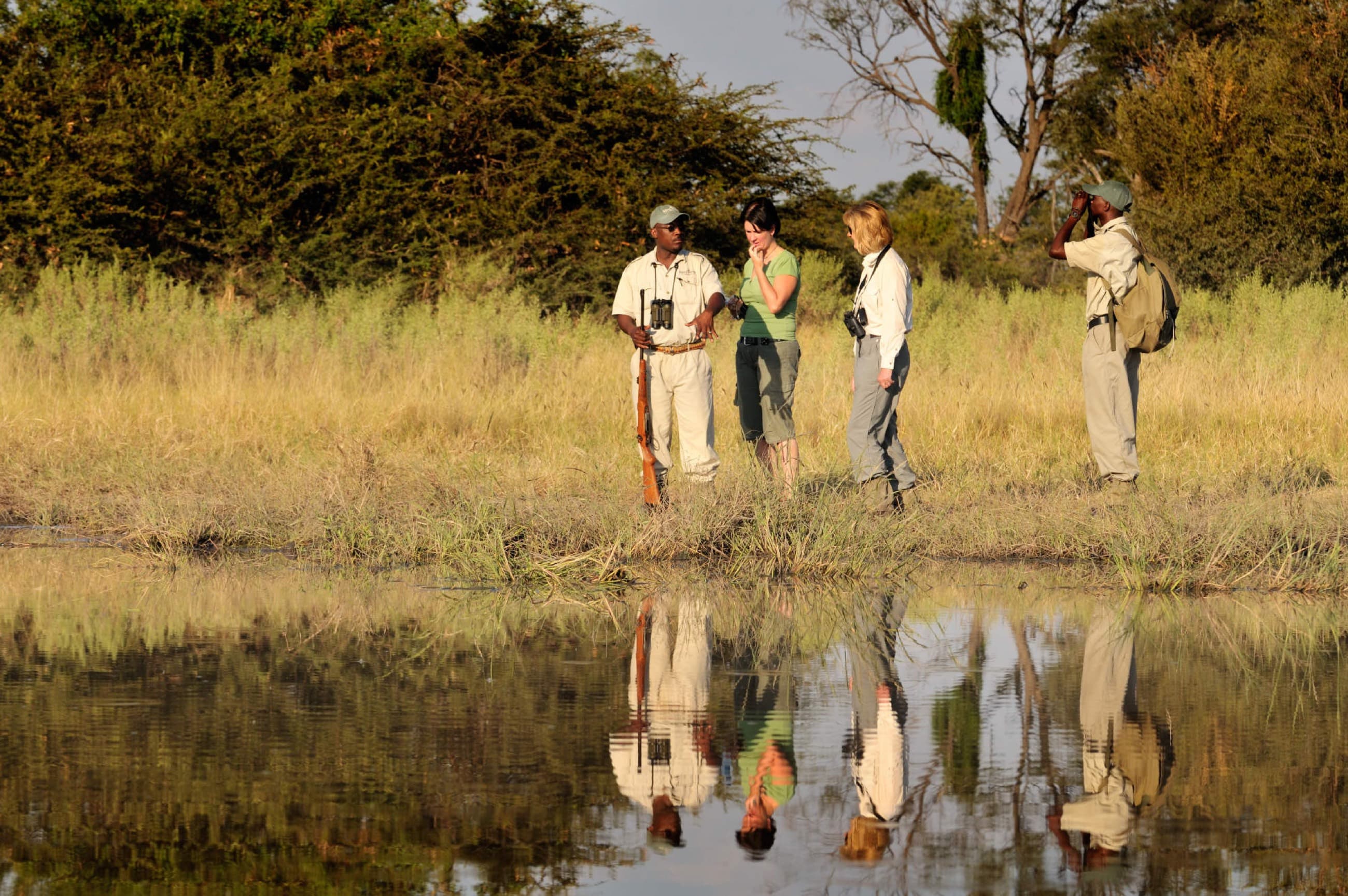 Walking safari guides and guests pausing beside a delta waterhole at golden hour